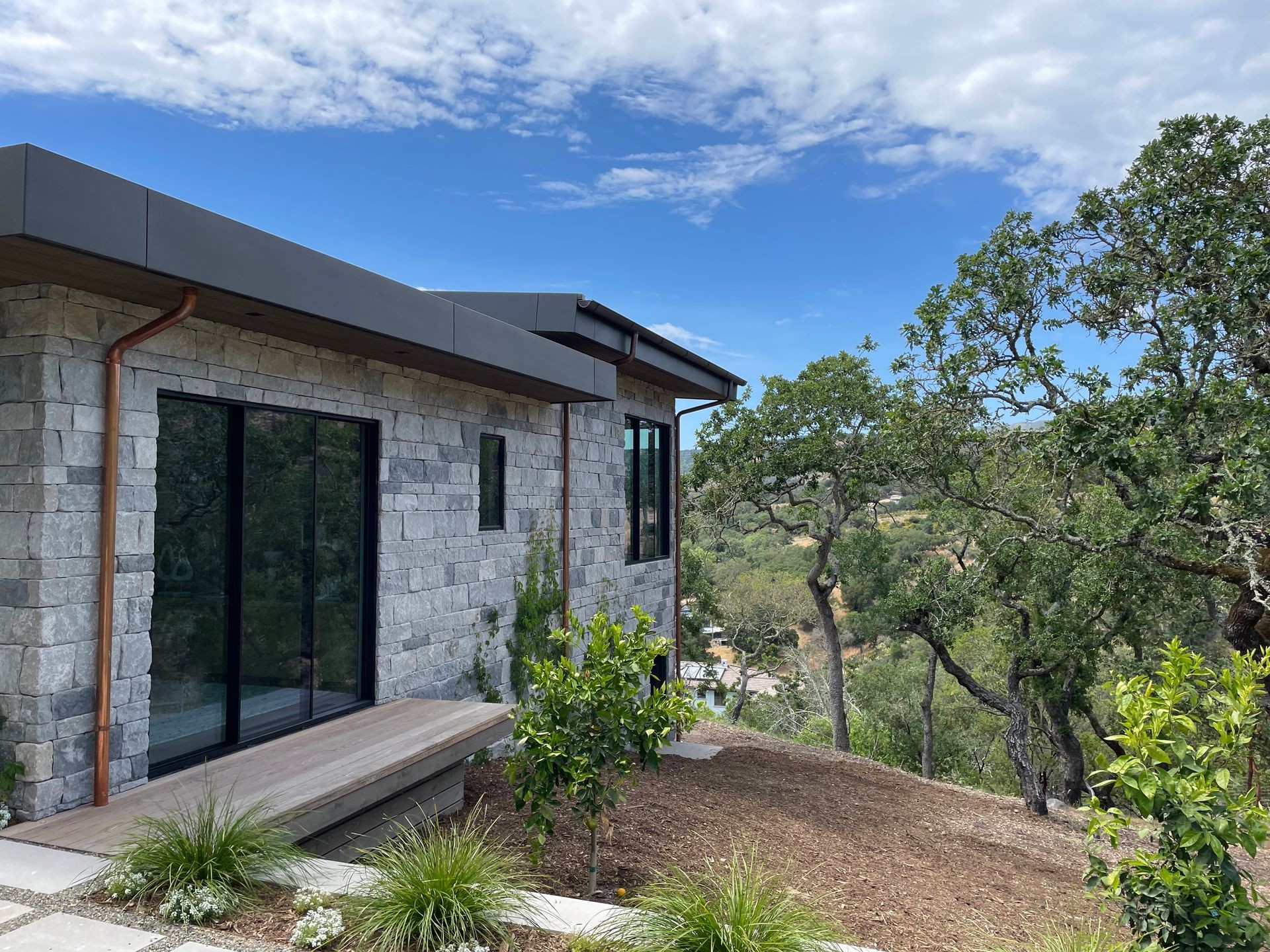 Modern stone house with large windows overlooking a lush green landscape on a sunny day.
