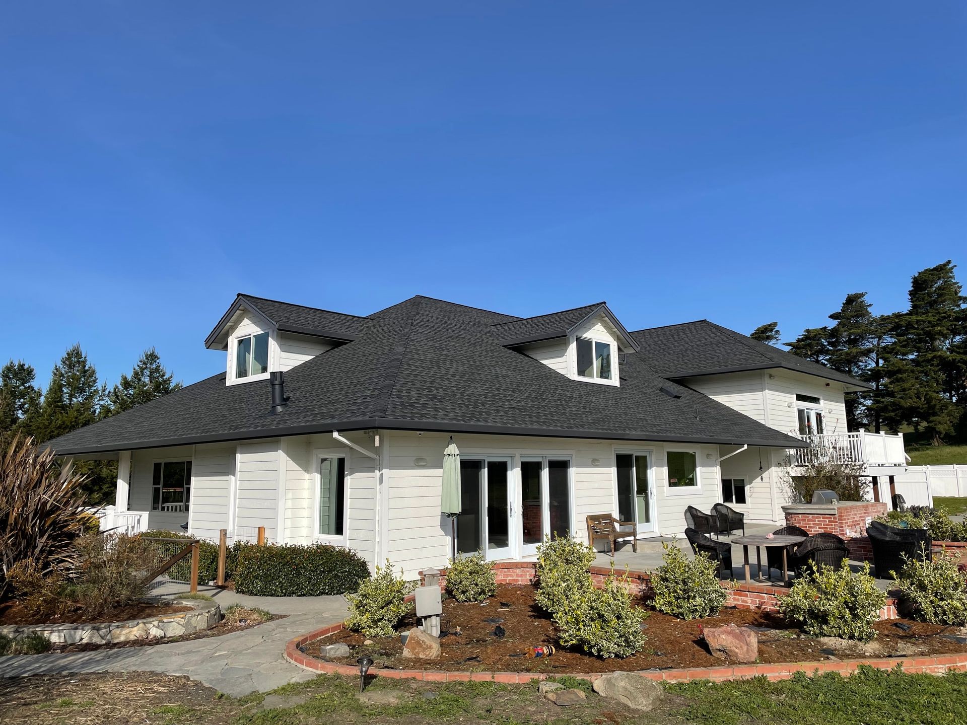 Two-story white house with dark roof, landscaped garden, and outdoor seating on a sunny day.