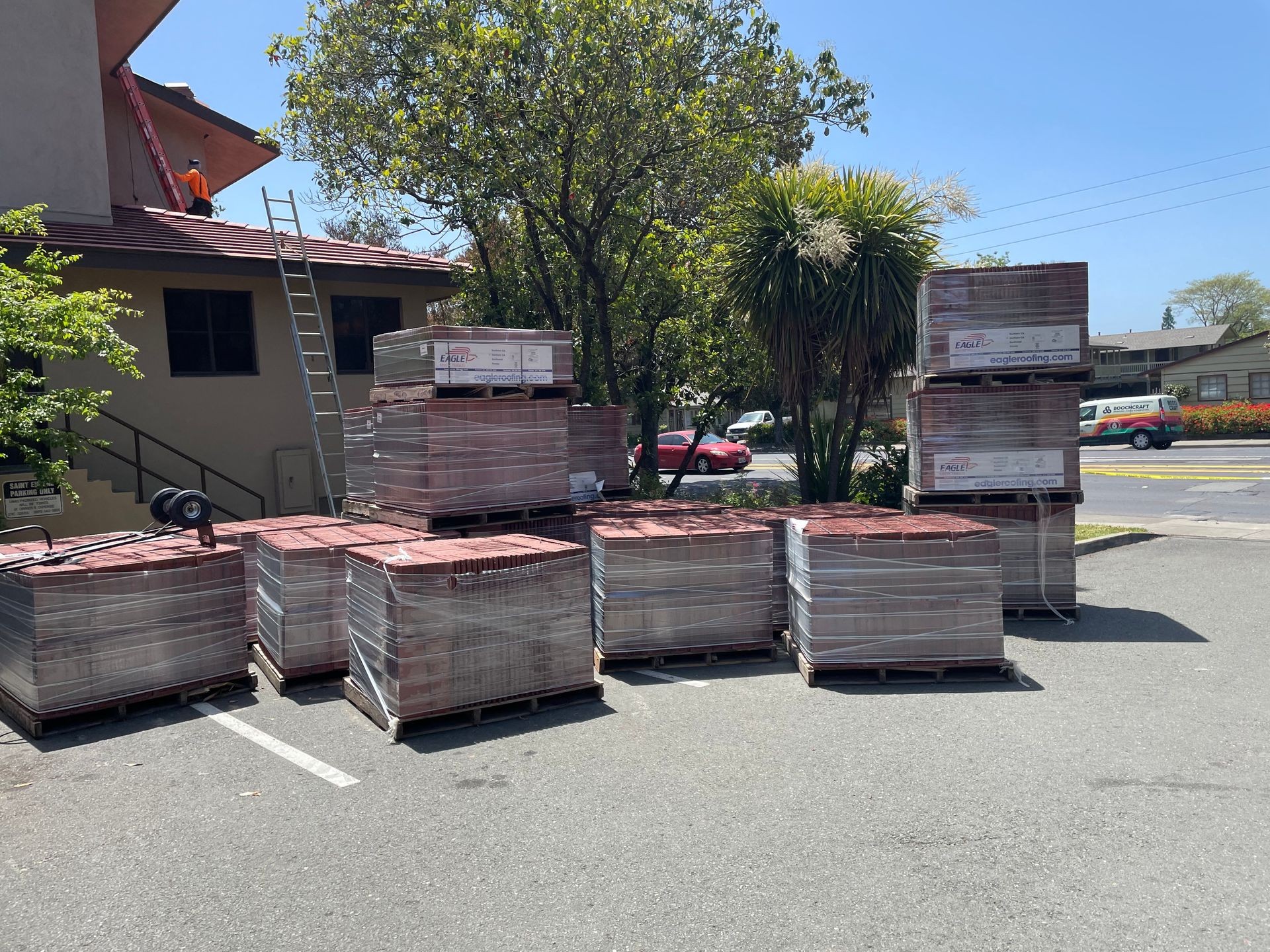 Pallets of roofing materials stacked outside a building with a ladder leaning against the roof.