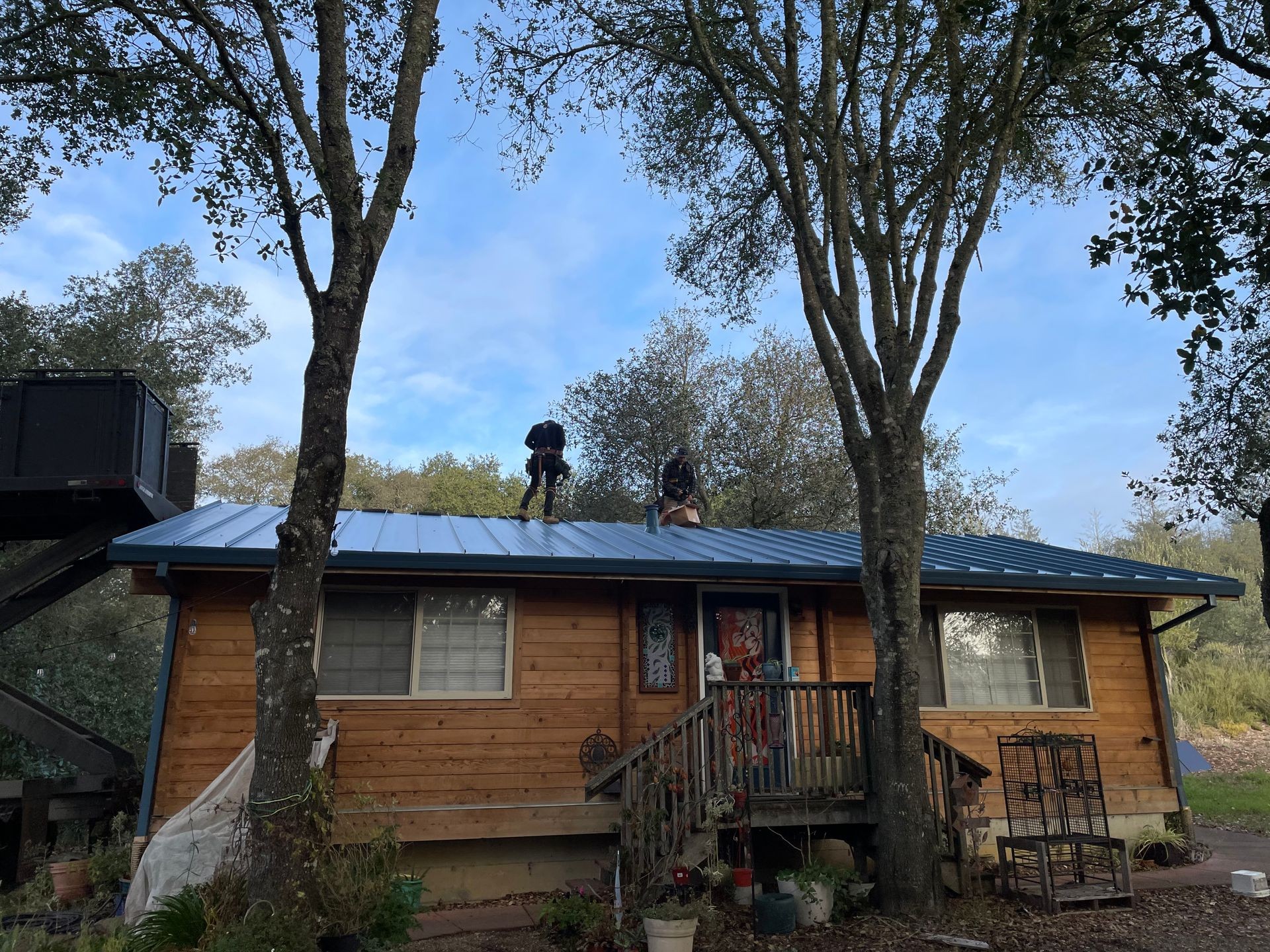 Two people working on the roof of a small wooden house surrounded by trees on a clear day.